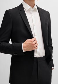 Zwarte op maat gemaakte blazer met notch revers, knoopmanchetten en een witte overhemden met kleine knopen en een opgevouwen kraag.