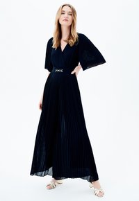 Mono negro con pliegues, escote en V, mangas anchas, cintura ajustada con herrajes metálicos y piernas sueltas y fluidas.