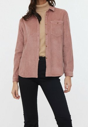 Kobieta ubrana w różową, zgaszoną koszulę ze sztruksu na guziki, pod którą ma beżowy golf, w połączeniu z czarnymi spodniami na neutralnym, białym tle.