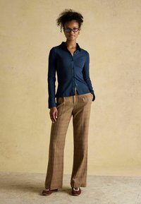 Navy ribbed button-up shirt paired with wide-leg brown plaid trousers. Model wears brown shoes. Background is a textured light color.