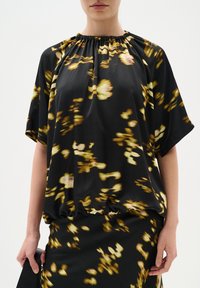 Blouse noire à manches courtes, avec un col froncé et des motifs floraux abstraits jaunes sur l'ensemble. Tissu doux et fluide.