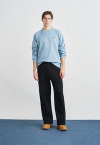 Νεαρό άτομο που στέκεται, φοράει ανοιχτό μπλε φούτερ, μαύρο παντελόνι και ταμπά μπότες, μπροστά σε απλό λευκό φόντο και μπλε πάτωμα.