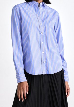 Kobieta ubrana w niebieską koszulę zapinaną na guziki z cienkimi białymi i różowymi pionowymi paskami oraz czarną plisowaną spódnicę.