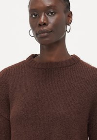Brauner Strickpullover mit rundem Ausschnitt, gerippter Struktur und lässiger Passform. Mit subtilen Details am Kragen akzentuiert.