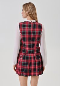 Abito senza maniche a quadri rossi e neri con vita aderente, gonna plissettata e camicia a maniche lunghe bianca sottostante, con chiusura sul retro.