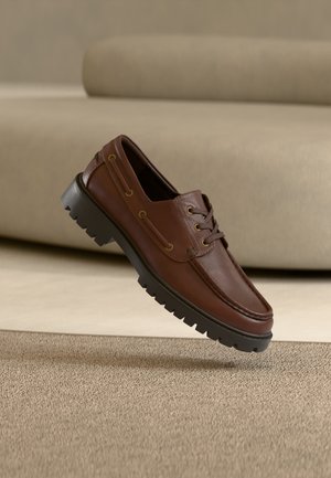 Hnedá kožená mokasína so šnurovaním a hrubou čiernou podrážkou s dezénom, ktorá je zavesená nad béžovým kobercom a pohovkou v pozadí.