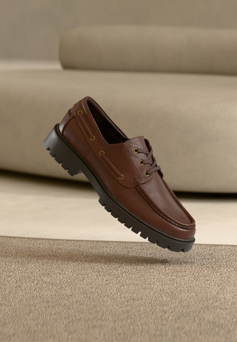 Mocasini maro din piele, cu șireturi, talpă groasă neagră cu profil, plutind deasupra covorului bej și a fundalului cu canapea.