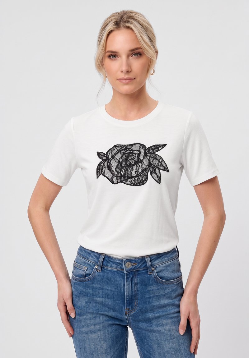 Femme blonde aux cheveux en ondulations lâches porte un t-shirt blanc avec un motif floral en dentelle noire et un jean bleu, debout avec une main sur la hanche.