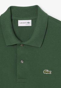Vihreä polo-paita, jossa on ribbireunus, kaksinappinen placket ja pieni brodeerattu krokotiili-logo vasemmalla puolella. Klassinen malli.
