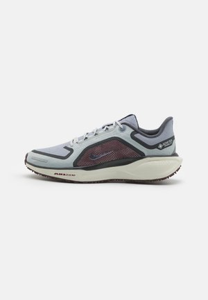 Graue und lila Laufschuhe mit einem Obermaterial aus Mesh, verstärkten Details und einer strukturierten Gummisohle mit sichtbarer Luftpolsterung.