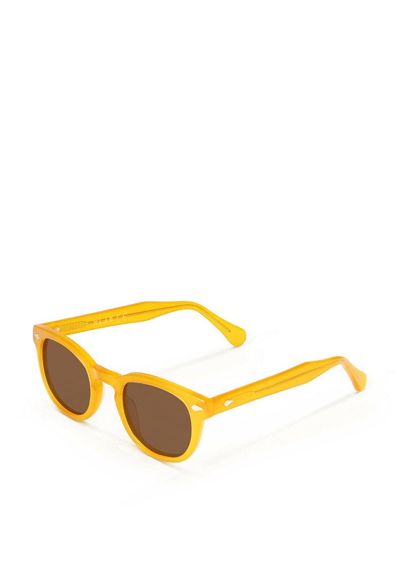 Gafas de sol amarillas con lentes de color marrón, forma de marco ovalado y patillas delgadas que presentan pequeños acentos en forma de diamante.