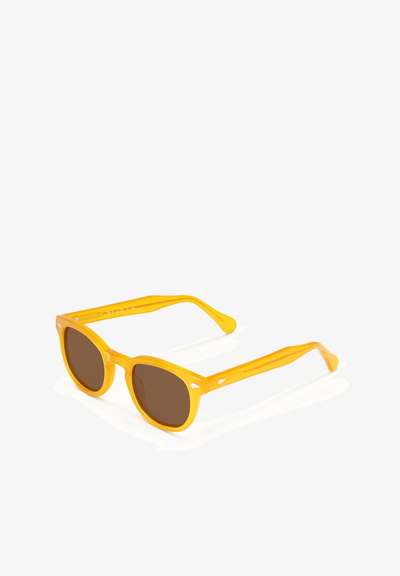 Gafas de sol amarillas con lentes de color marrón, forma de marco ovalado y patillas delgadas que presentan pequeños acentos en forma de diamante.