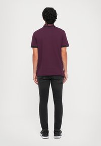 Homem com cabelo encaracolado está de costas, vestindo uma polo roxa de mangas curtas, jeans slim pretos e ténis pretos com solas brancas.