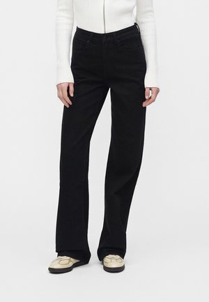 Personne portant un pantalon large noir, un haut blanc côtelé à manches longues avec fermeture éclair et des chaussures beiges, debout devant un fond uni.