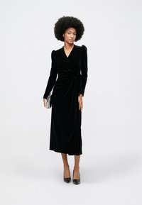 Ženska z afro frizuro nosi dolgo črno žametno obleko z zavojem in črne pete, v roki pa drži svetlo obarvano torbico.