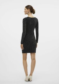 Vestido negro con mangas largas, patrón texturizado en zigzag, silueta ajustada y longitud por encima de la rodilla. Llevado con tacones beige.