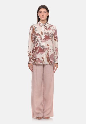 Donna che indossa una camicia a maniche lunghe con motivo nelle tonalità del beige e marrone, abbinata a pantaloni ampi rosa chiaro e sandali con punta aperta.