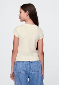 Mergina su ilgais rudos spalvos plaukais vilkinti šviesiai geltoną trumpomis rankovėmis palaidinę ir mėlynas džinsines kelnes, žiūrinti nuošaly priešais paprastą foną.