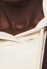 Haut à capuche de couleur crème avec cordon de serrage, présentant un tissu texturé et un accent marron sur un côté, vue en gros plan du col.