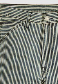 Pantalones de mezclilla a rayas con rayas verticales en azul y crema, que cuentan con un botón de metal y detalles cosidos en los bolsillos y la cinturilla.