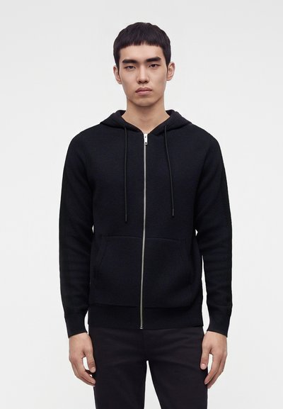 Young man with short black hair wearing a black zip-up hoodie and black pants standing against a plain light background.