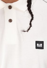 Hvit polo-skjorte med teksturert stoff, med tre brune knapper og et svart rektangulært merke med "WEEKEND OFFENDER"-etikett.