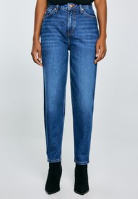 Jeans z wysokim stanem w niebieskim kolorze, proste nogawki, z wyblakłym odcieniem, widocznymi szwami i przednimi kieszeniami, noszone z czarnymi botkami za kostkę.