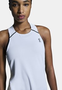 Top deportivo blanco sin mangas con una tela texturizada, que presenta detalles negros y un escote redondo. Logotipo mostrado en el lado frontal.