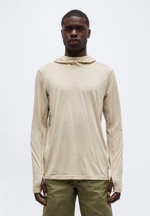 Giovane uomo con indosso una felpa beige con cappuccio a maniche lunghe e pantaloni verde oliva, in piedi contro uno sfondo chiaro e uniforme.