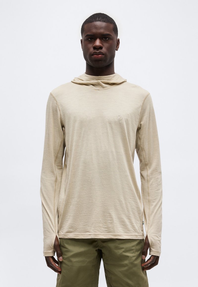 Noormees, kes kannab beežikat pika varrukaga kapuutsiga särki ja oliivirohelisi pükse, seisab ühevärvilise hele tausta ees.