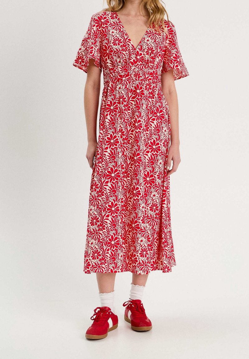 Robe midi rouge à fleurs avec des manches courtes volantées, un décolleté en V et des motifs de fleurs blanches. Portée avec des baskets rouges et des chaussettes blanches à la cheville.