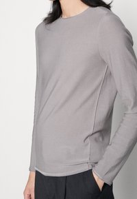 Szara koszula z długim rękawem wykonana z miękkiego materiału, z okrągłym dekoltem, szwami bocznymi i luźnym krojem. Dodatkowe detale to przeszycia.