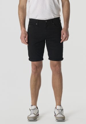 Hombre vistiendo pantalones cortos negros hasta la rodilla con dobladillo, zapatillas blancas con detalles grises y camiseta blanca sobre un fondo liso.