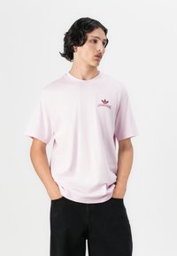 Junger Mann mit schwarzen lockigen Haaren, der ein hellrosa Adidas T-Shirt und schwarze Hose trägt, die Hände in den Taschen, und zur Seite schaut.