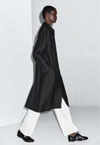 Cappotto lungo nero con design dritto, tasche laterali e bottoni; indossato sopra pantaloni bianchi a gamba larga e scarpe nere stringate.