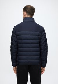 Sötétkék párnázott kabát, melynek texturált felső része és sima alsó része van. Álló gallérral és vízszintes varrással rendelkezik a háton.