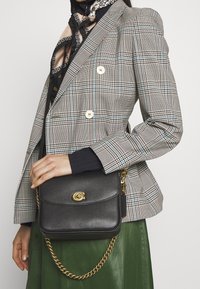 Borsa a tracolla in pelle nera con catena dorata, dotata di una patta frontale e di un logo decorativo. Il modello indossa un blazer scozzese e una gonna verde.