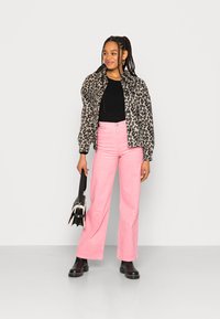 Casaco bomber com padrão de leopardo, top preto canelado, calças largas em veludo cotelê cor-de-rosa, botas de tornozelo pretas, segurando uma pequena mala preta.
