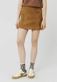 Mini-jupe en daim marron avec deux boutons noirs, accompagnée de chaussettes à motif et de baskets, mettant en valeur une tenue décontractée et superposée.