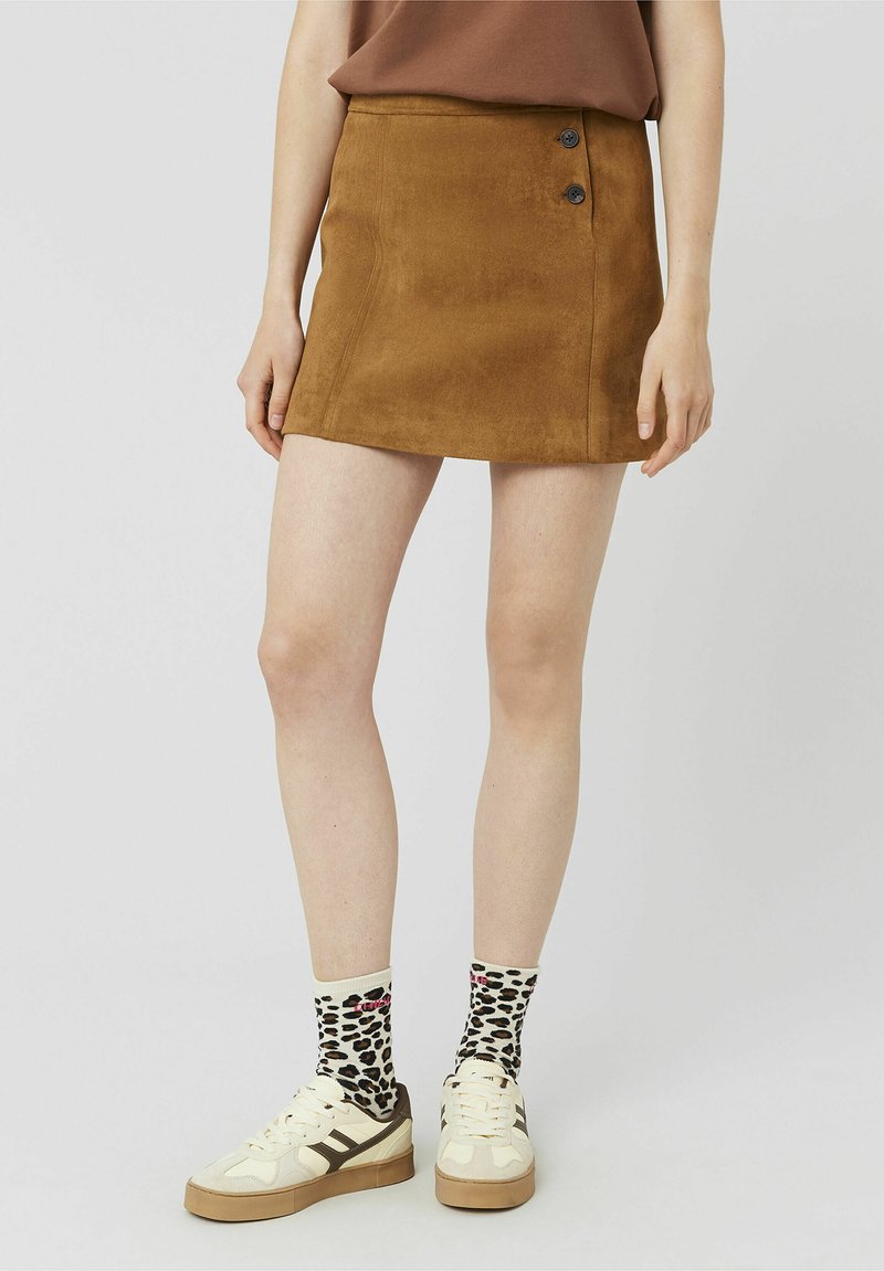 Mini-jupe en daim marron avec deux boutons noirs, accompagnée de chaussettes à motif et de baskets, mettant en valeur une tenue décontractée et superposée.