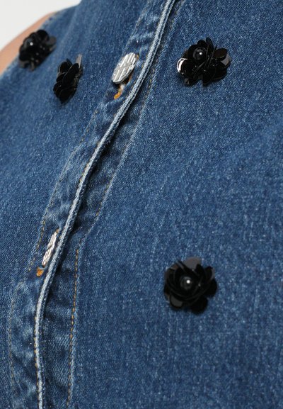 Denim tkanina z modrim odtenkom, ki ima srebrne in črne cvetlične okraske po sprednji strani, poudarjene z kontrastnim šivanjem.