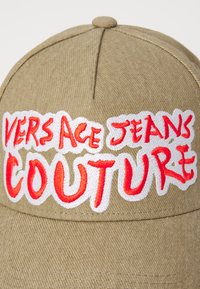 Casquette beige avec le texte brodé "VERSACE JEANS COUTURE" en blanc et rouge. Dotée d'un peak structuré et d'un tissu texturé.
