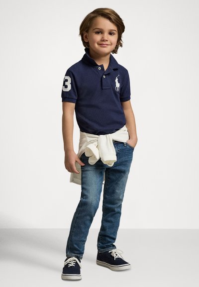 Tmavomodré polo tričko s bielym akcentom na golieri a číslom 3. Svetlomodré džínsy a biely sveter zviazaný okolo pása. Tmavomodré topánky.