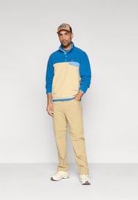 Μπλε και μπεζ φλις πουλόβερ με γιακά με κουμπιά και τσέπη. Καφέ παντελόνι convertible. Άσπρα αθλητικά παπούτσια. Καπέλο με λογότυπο.