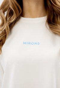 Bluza z okrągłym dekoltem w miękkim białym materiale z jasnoniebieskim haftem z napisem "MIRONS" umieszczonym na środku klatki piersiowej.