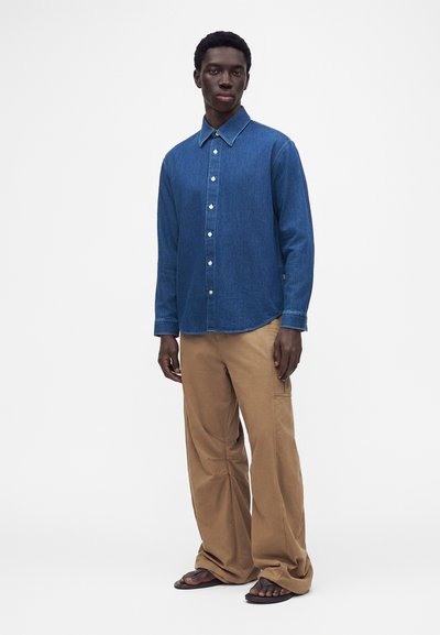 Homme debout portant une chemise bleue à manches longues boutonnée, un pantalon beige ample et des sandales foncées sur fond blanc.
