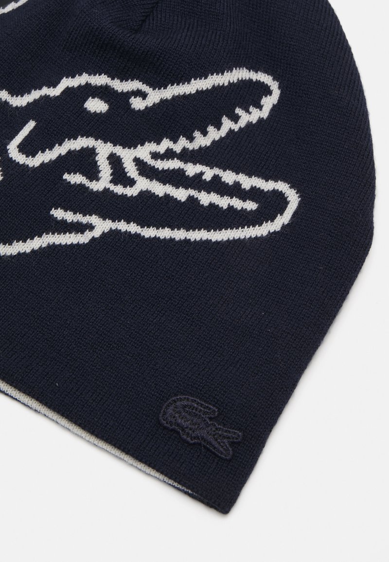 Bonnet tricot navy mettant en avant un logo de crocodile blanc bien visible, avec un léger détail de marque brodé en bas.