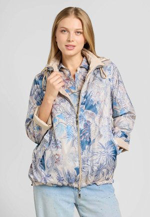 Donna che indossa una giacca chiara con motivo floreale e cerniera frontale e jeans azzurri con piccole decorazioni.