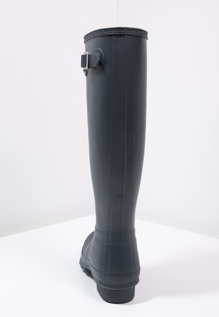 Bota de goma negra hasta la rodilla con un acabado texturizado, correa ajustable en el tobillo y suela robusta, mostrada con la parte trasera hacia adelante.