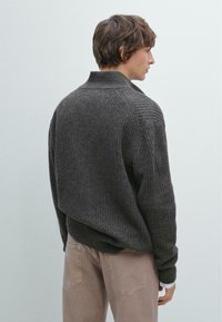 Mørkegrå ribbet sweater med høj krave og sænkede skuldre, båret over lyse beige bukser, der viser en afslappet pasform og tekstureret mønster.